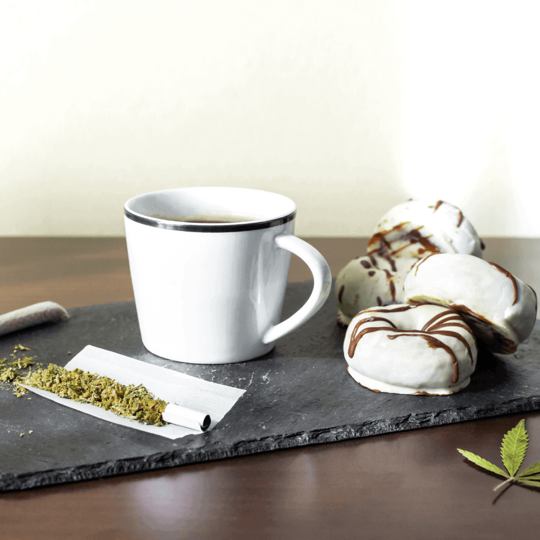 an unrolled joint sits next to a mug of coffee and frosted donuts, the perfect snack for the munchies